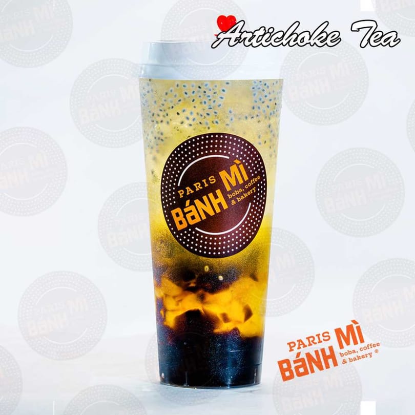 A clear plastic cup of Paris Banh Mi Artichoke Tea with dark and light layers, and seeds.