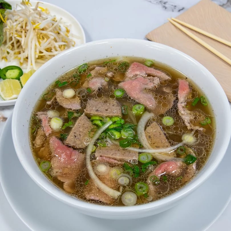 A bowl of Vietnamese pho with beef, meatballs, green onions, and bean sprouts on the side.