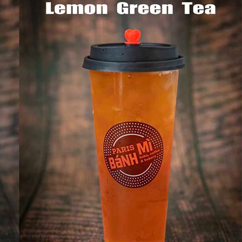 A tall plastic cup of Paris Banh Mi Lemon Green Tea with a black lid and red heart stopper.