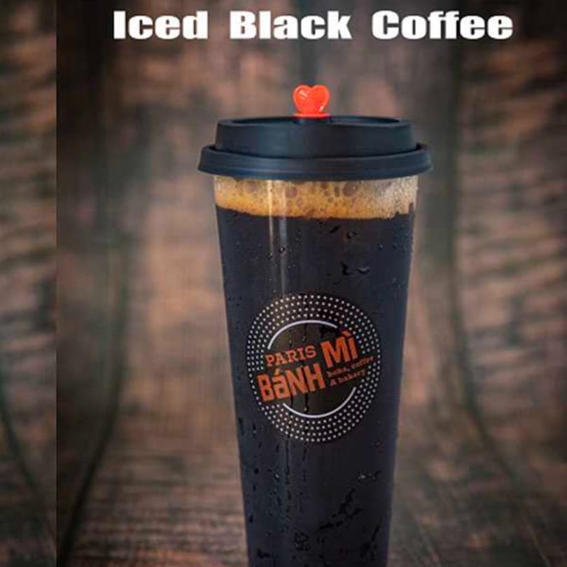 Iced black coffee in a clear cup with a black lid and red heart stopper, on a wooden background.
