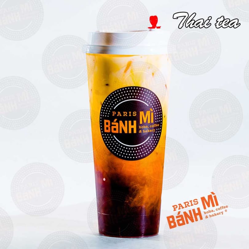 A tall plastic cup of Thai tea with a Paris Banh Mi logo, showing layers of dark tea and creamy milk.