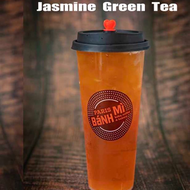 A tall plastic cup of Jasmine Green Tea with a black lid and red heart stopper, on a wooden surface.