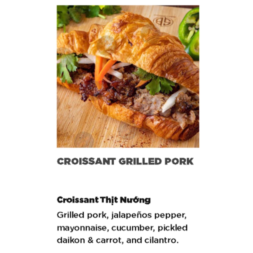 Grilled pork croissant sandwich with pickled daikon, carrots, and cilantro on a wooden board.