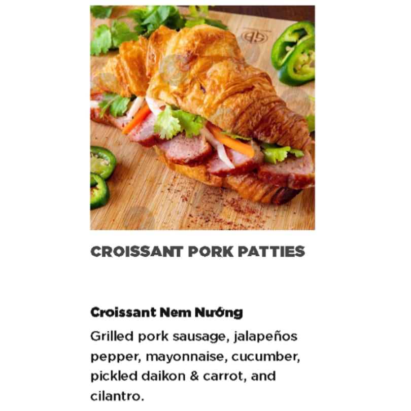 A Vietnamese Croissant Nem Nuong with grilled pork sausage, vegetables, and cilantro on a wooden board.