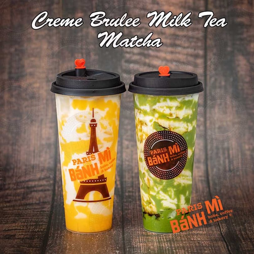 Two Paris Banh Mi Creme Brulee Milk Teas, one yellow and one green matcha, on a wooden surface.