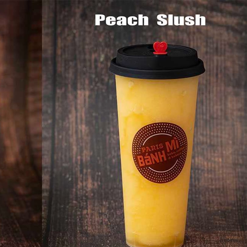 A Paris Banh Mi peach slush in a clear cup with a black lid and red heart stopper.