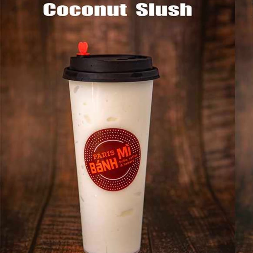 A Paris Banh Mi Coconut Slush in a clear cup with a red heart stopper on a wooden background.