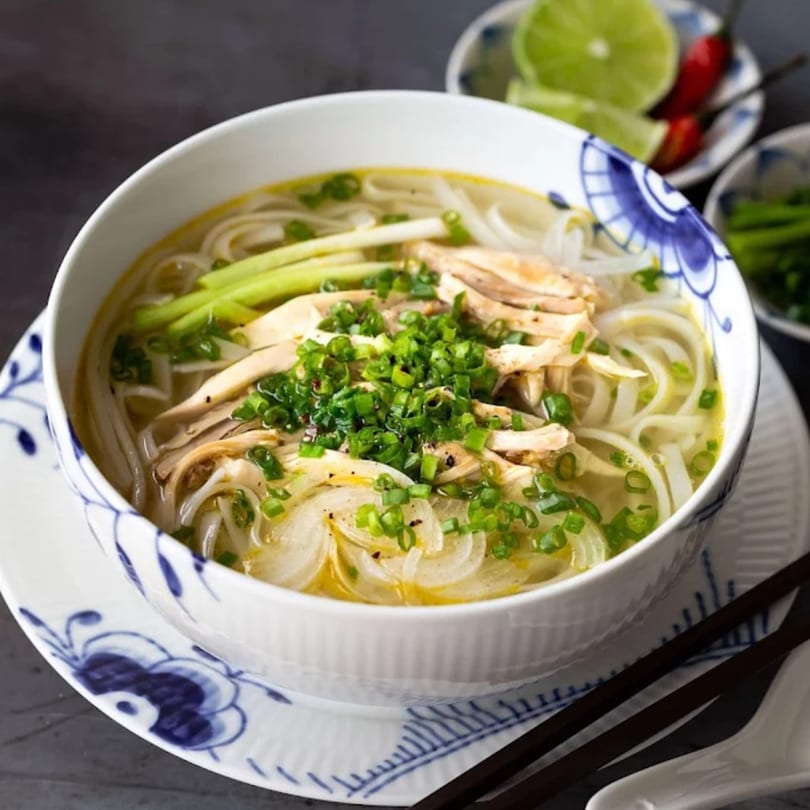 A bowl of Vietnamese chicken pho with noodles, green onions, and lime wedges.