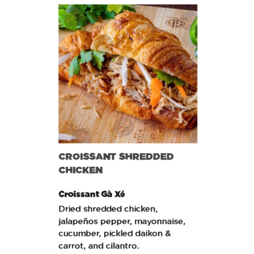 A Croissant Gà Xé, filled with shredded chicken, pickled daikon, carrots, and cilantro on a wooden board.