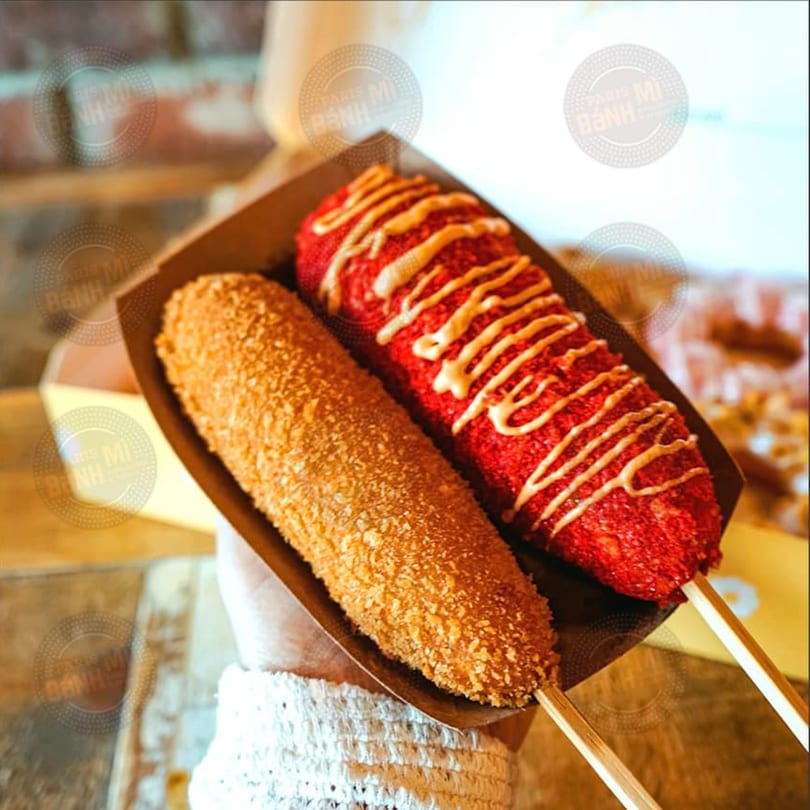 Two corn dogs, one golden brown and one red with white sauce, held in a paper tray.