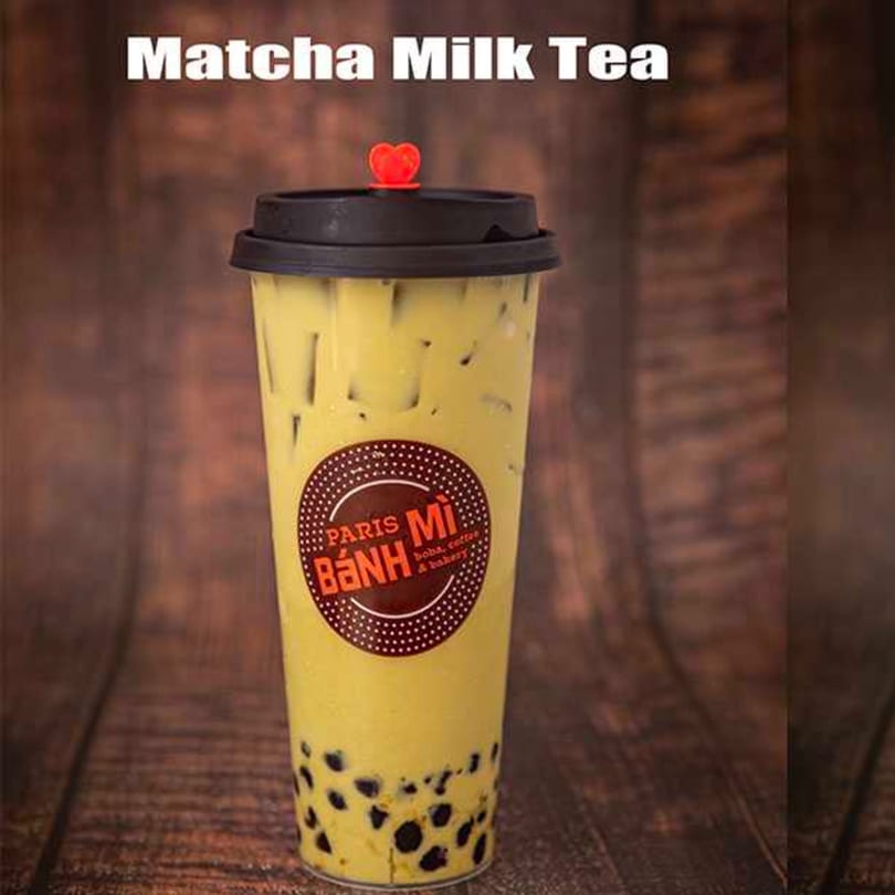 A tall plastic cup of Paris Banh Mi matcha milk tea with boba, on a wooden surface.