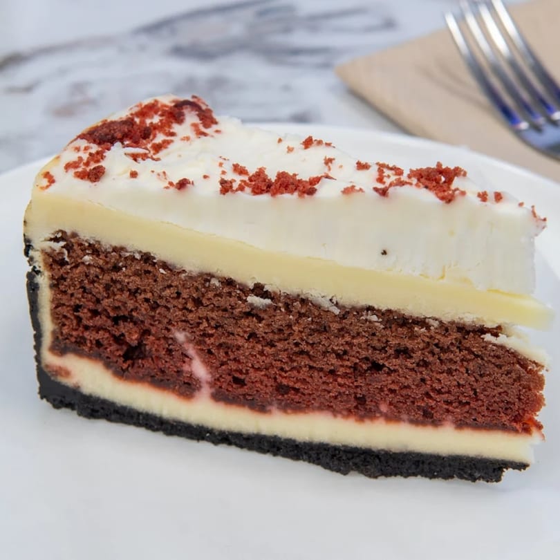 A slice of red velvet cheesecake with white frosting and red crumbs on a white plate.