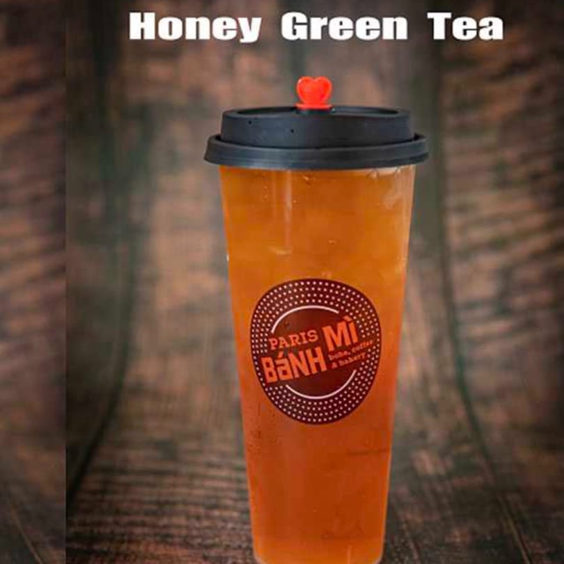 Honey Green Tea in a clear plastic cup with a black lid and red heart stopper, on a wooden surface.