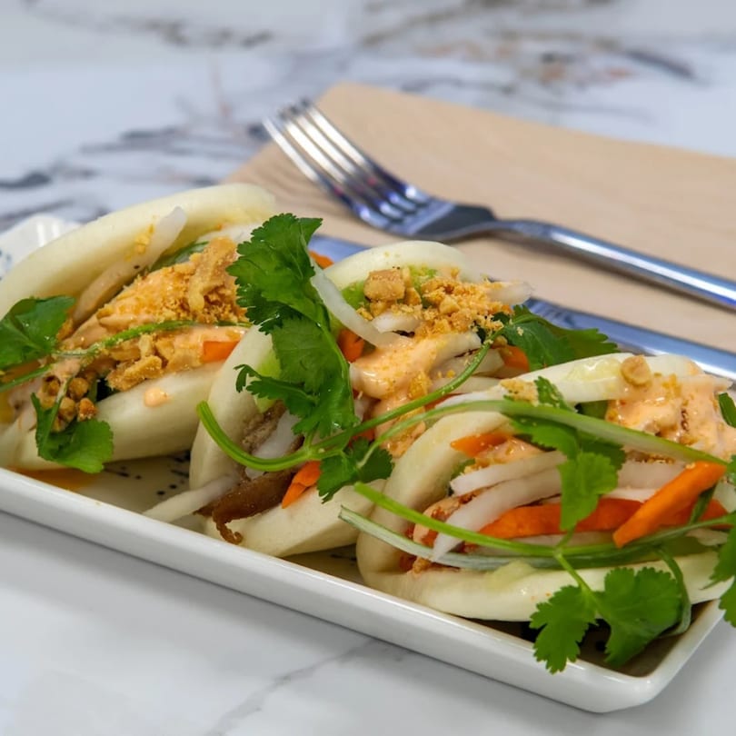 Three Vietnamese bao buns filled with meat, vegetables, sauce, and cilantro on a white plate.